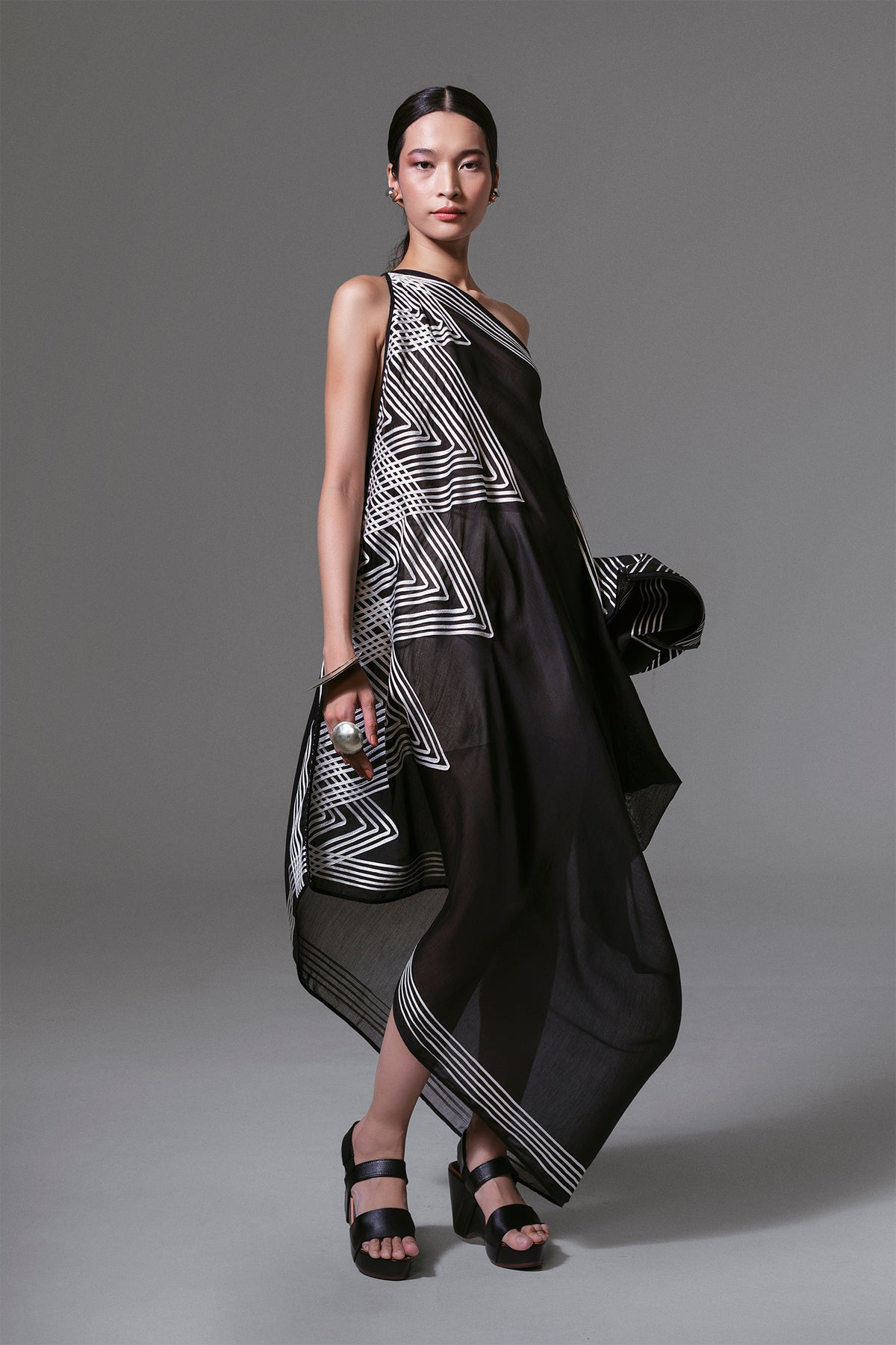 Model wearing black one-shoulder asymmetrical dress with white geometric ribbon appliqué and sheer silk-cotton finish.