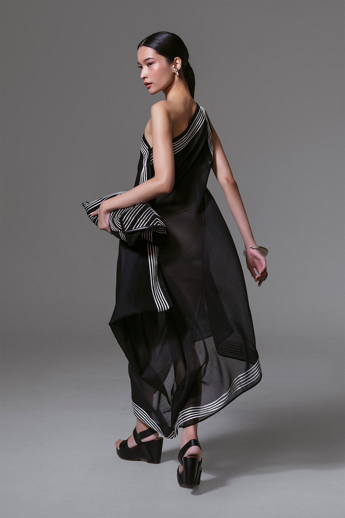 Model wearing black one-shoulder asymmetrical dress with white geometric ribbon appliqué and sheer silk-cotton finish.