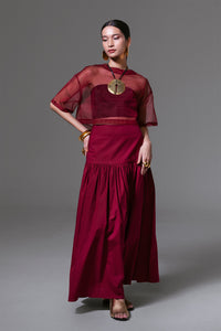 Model wears a bordeaux cotton maxi skirt with a fitted yoke and gathered wide hem, paired with a sheer cropped top.