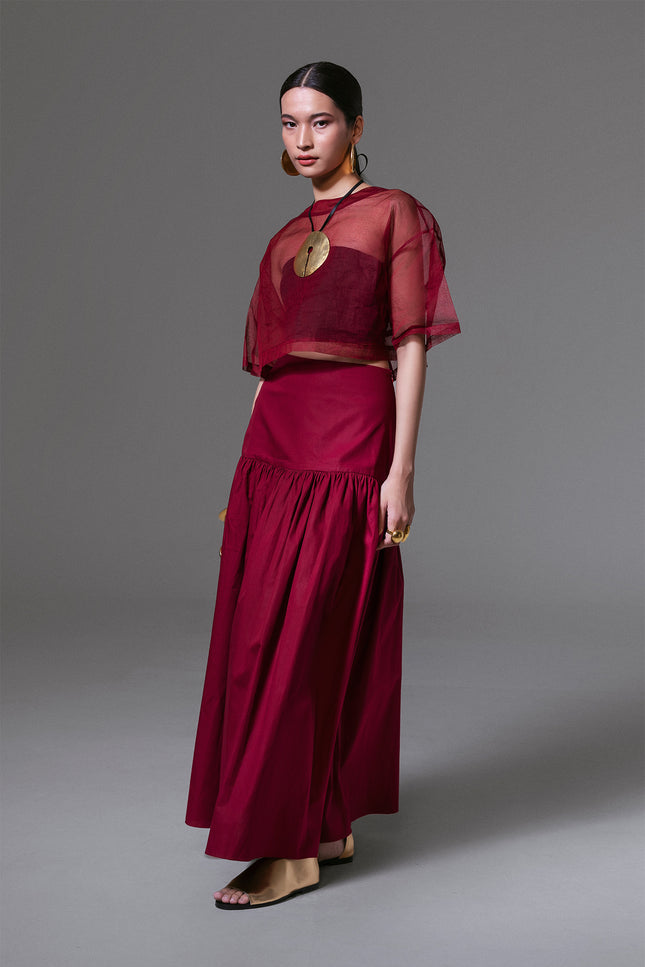 Model wears a bordeaux cotton maxi skirt with a fitted yoke and gathered wide hem, paired with a sheer cropped top.