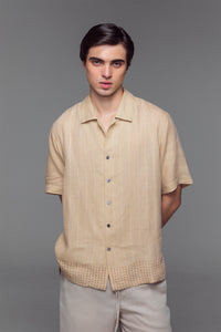 Model wearing stone-colored linen camp shirt with open collar, button front, and dual-colour hand-stitched detailing.