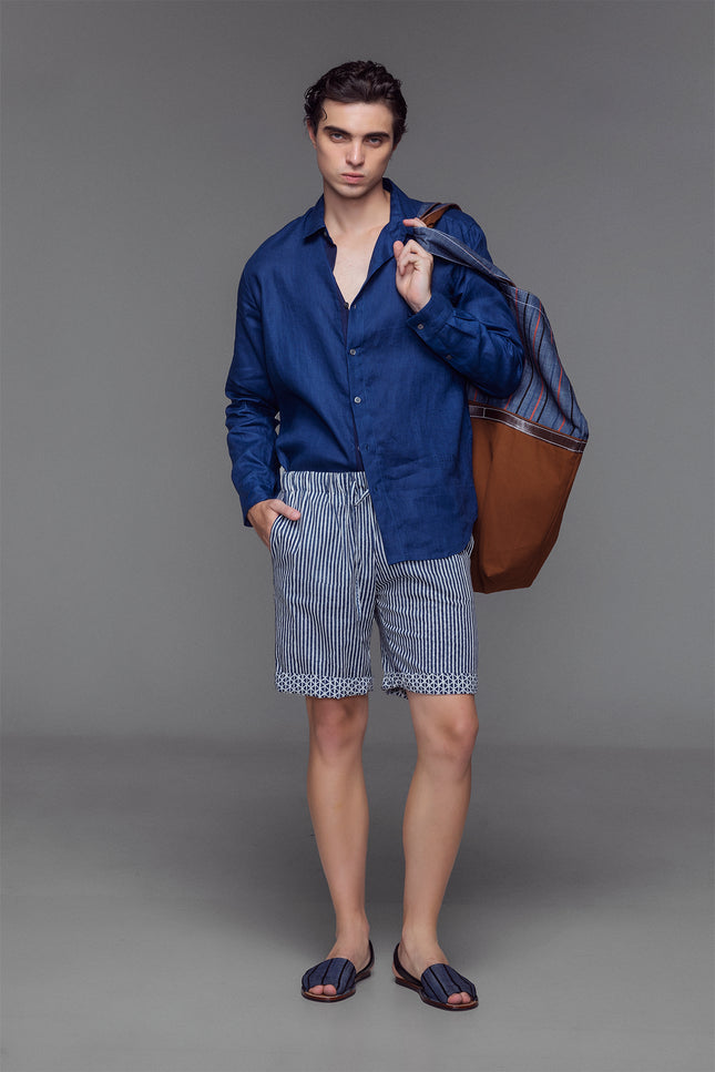 Model wearing reversible Mical Shorts in oriental blue and off-white blom print, featuring an elastic waistband with drawstrings, side and back pokets, and a short, breathable cotton silhouette for warm-weather wear.