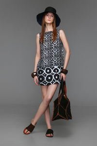 Model wears sleeveless black and off-white linen dress featuring bold circular prints, banded hem, boat neckline, and cotton lining.