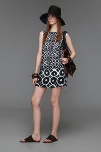 Model wears sleeveless black and off-white linen dress featuring bold circular prints, banded hem, boat neckline, and cotton lining.