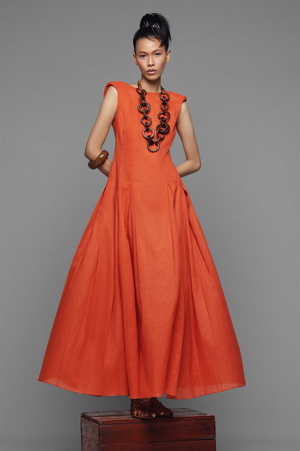 Model wearing Nadia Dress in an A-line silhouette with structured shoulders, boat neckline, and deep V back. Pleated detailing flows from the hip, crafted in a breathable ramie-cotton blend for refined summer occasions.