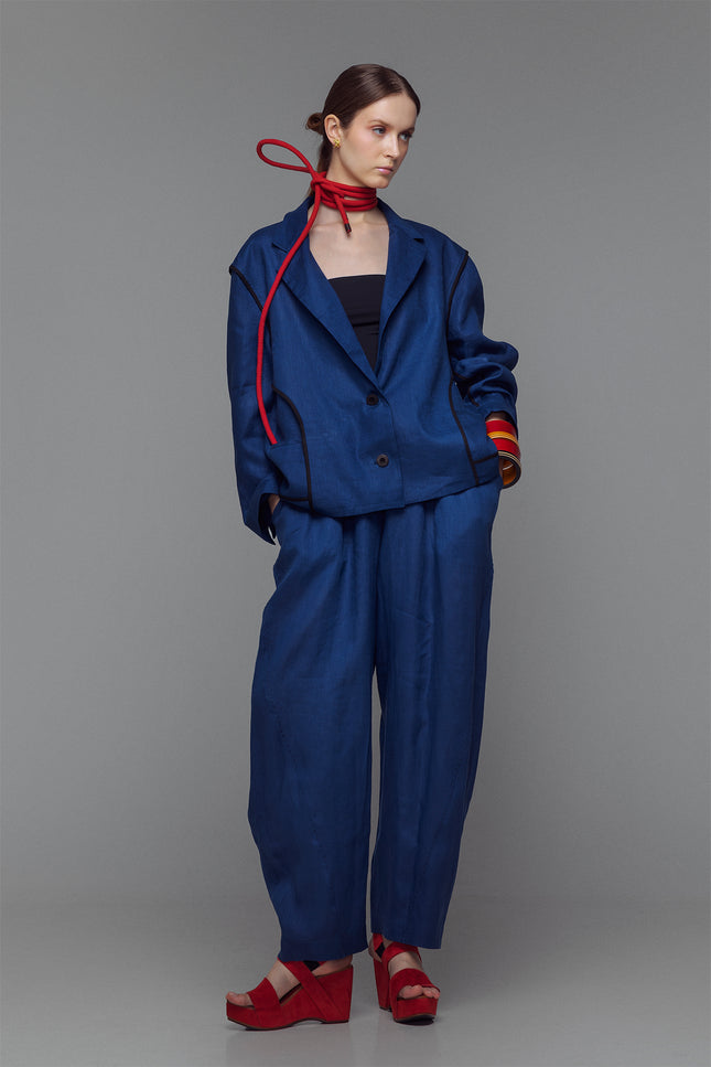 Model wearing indigo balloon pants with hand-stitched accents and an elastic waist, styled with a matching jacket and red sandals.