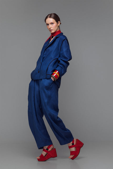 Model wearing indigo balloon pants with hand-stitched accents and an elastic waist, styled with a matching jacket and red sandals.