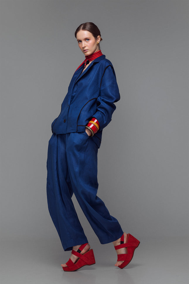 Model wearing indigo balloon pants with hand-stitched accents and an elastic waist, styled with a matching jacket and red sandals.