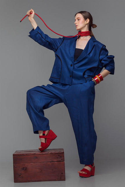 Model wearing indigo balloon pants with hand-stitched accents and an elastic waist, styled with a matching jacket and red sandals.