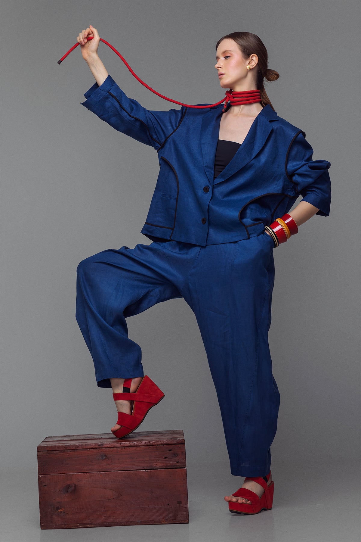Model wearing indigo balloon pants with hand-stitched accents and an elastic waist, styled with a matching jacket and red sandals.