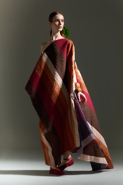 Model wearing the Oreta Dress with an asymmetrical draped silhouette, one-shoulder design, multicolored diagonal appliqué stripes, adjustable side buttons, and a side slit.