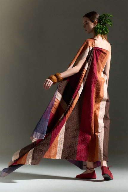 Model wearing the Oreta Dress with an asymmetrical draped silhouette, one-shoulder design, multicolored diagonal appliqué stripes, adjustable side buttons, and a side slit.