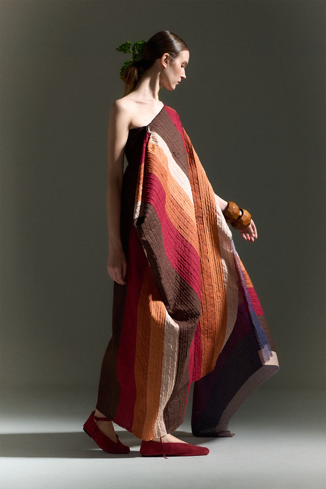 Model wearing the Oreta Dress with an asymmetrical draped silhouette, one-shoulder design, multicolored diagonal appliqué stripes, adjustable side buttons, and a side slit.