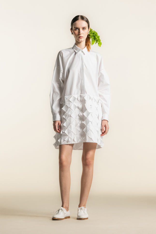 Model wearing the Nann Shirt Dress with a relaxed tailored silhouette, dropped shoulders, and long cuffed sleeves, featuring a contrasting three dimensional geometric panel on the lower body in lightweight white cotton.