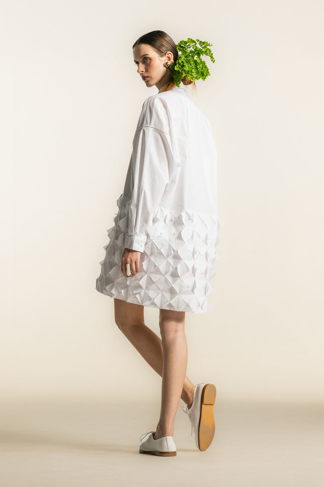 Model wearing the Nann Shirt Dress with a relaxed tailored silhouette, dropped shoulders, and long cuffed sleeves, featuring a contrasting three dimensional geometric panel on the lower body in lightweight white cotton.